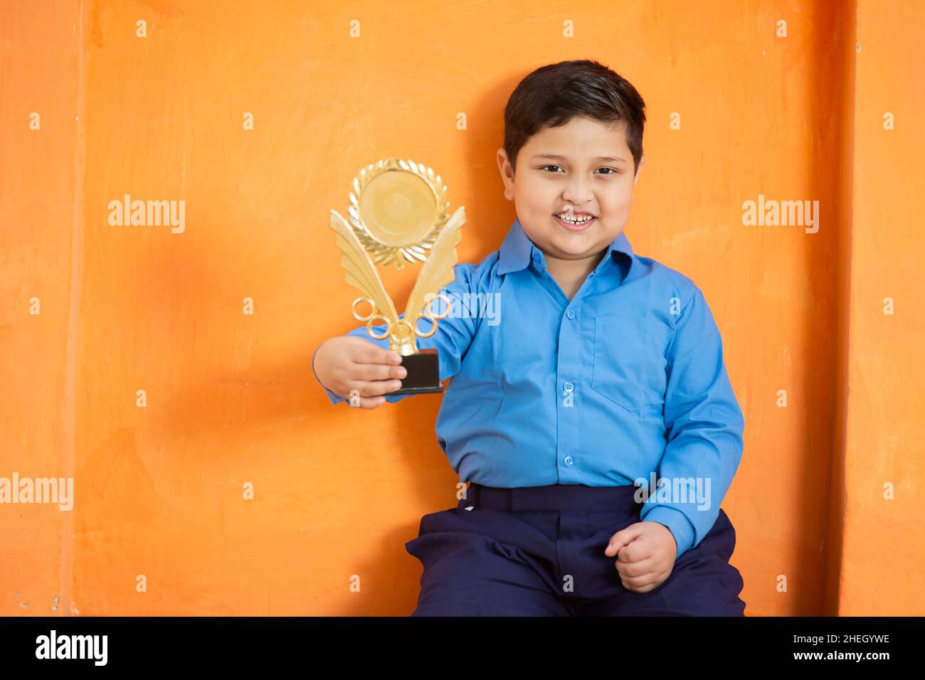 Portrait of happy adorable cute indian boy in school uniform showing trophy or celebrating victory,Cheerful male kid holding winning prize against ora Stock Photo