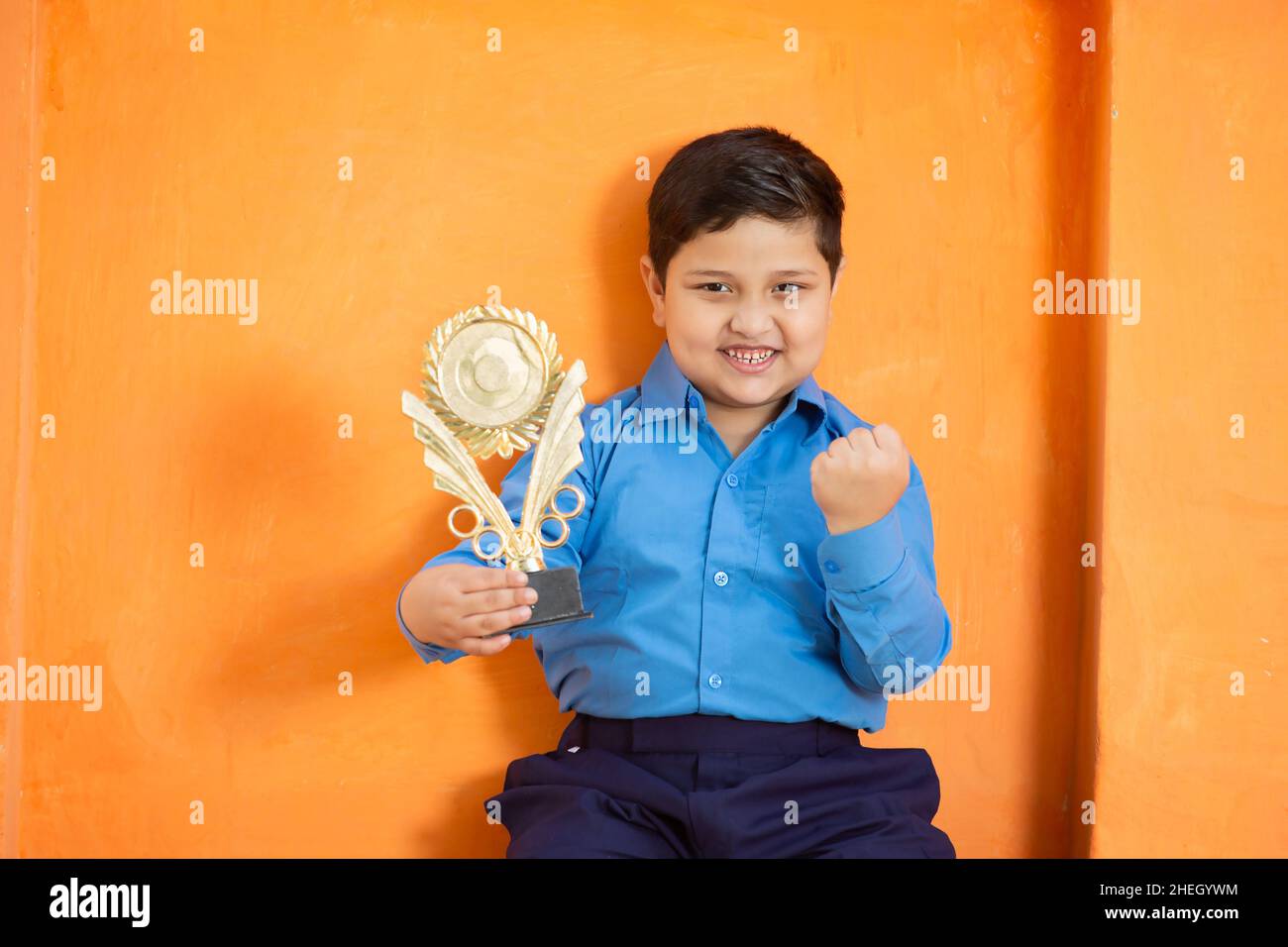 Happy adorable cute indian boy in school uniform celebrating victory with trophy,Cheerful excited male kid holding winning prize against orange backgr Stock Photo