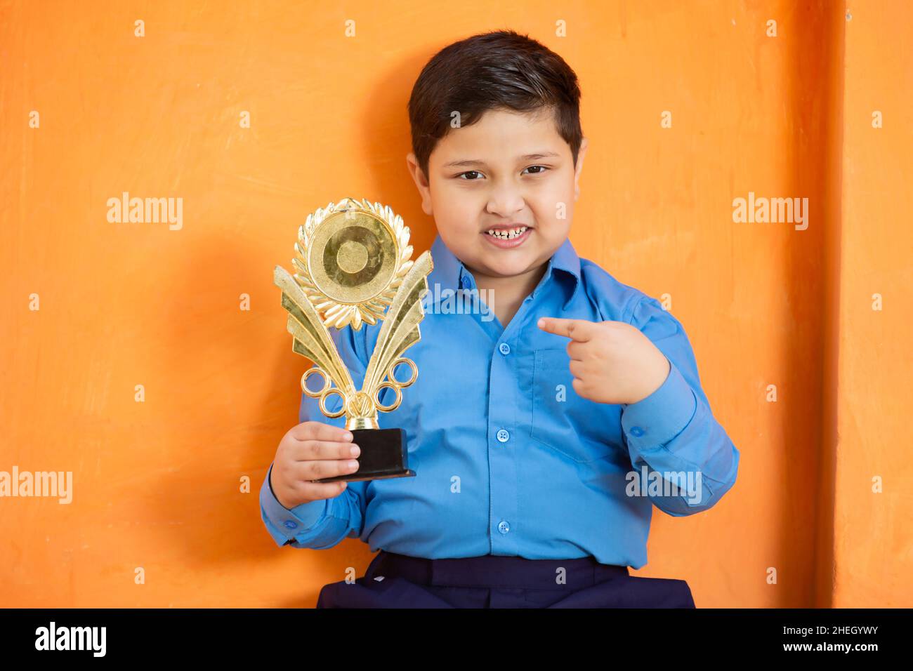 Portrait of happy adorable cute indian boy in school uniform celebrating victory with trophy,Cheerful male kid holding winning prize against orange ba Stock Photo
