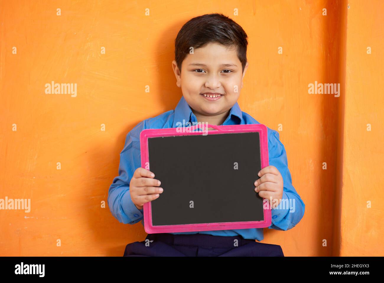 Portrait of happy cute little indian boy in school uniform holding blank slate against orange background, Adorable elementary kid showing black board. Stock Photo