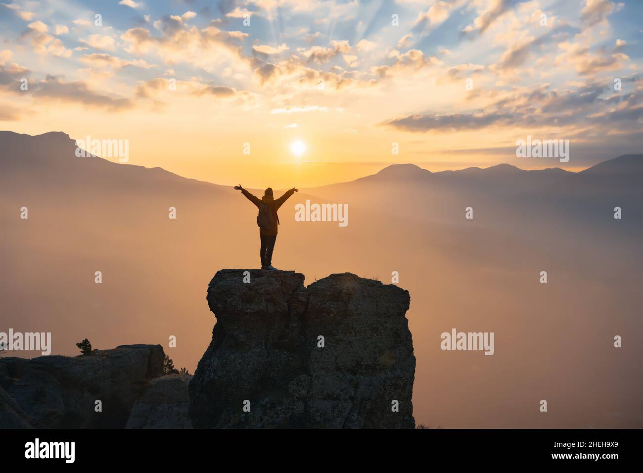 Silhouette of sporty girl standing on mountain top with raised arms at sunset Stock Photo