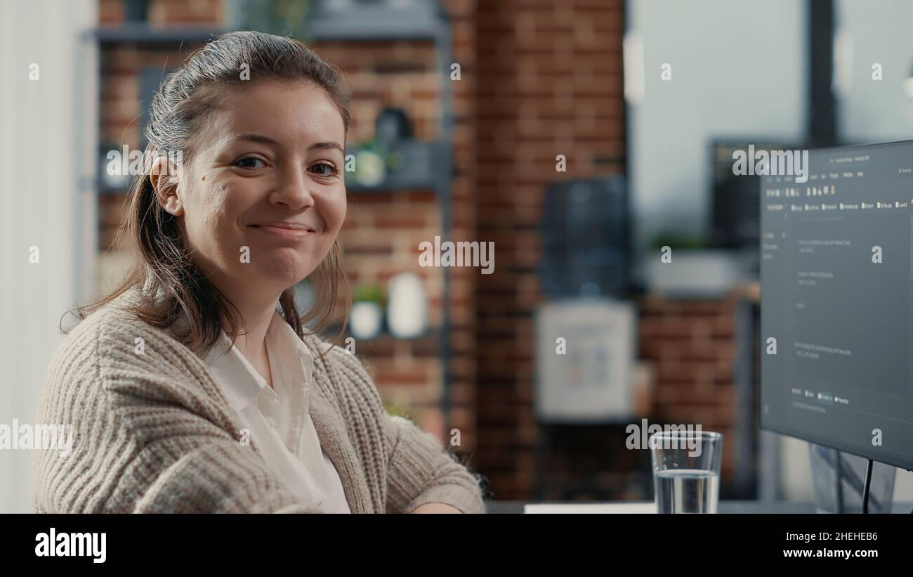 Portrait of software developer typing programming language on computer keyboard turning head and smiling at camera. Casual database admin sitting at desk computing big data feeling confident. Stock Photo