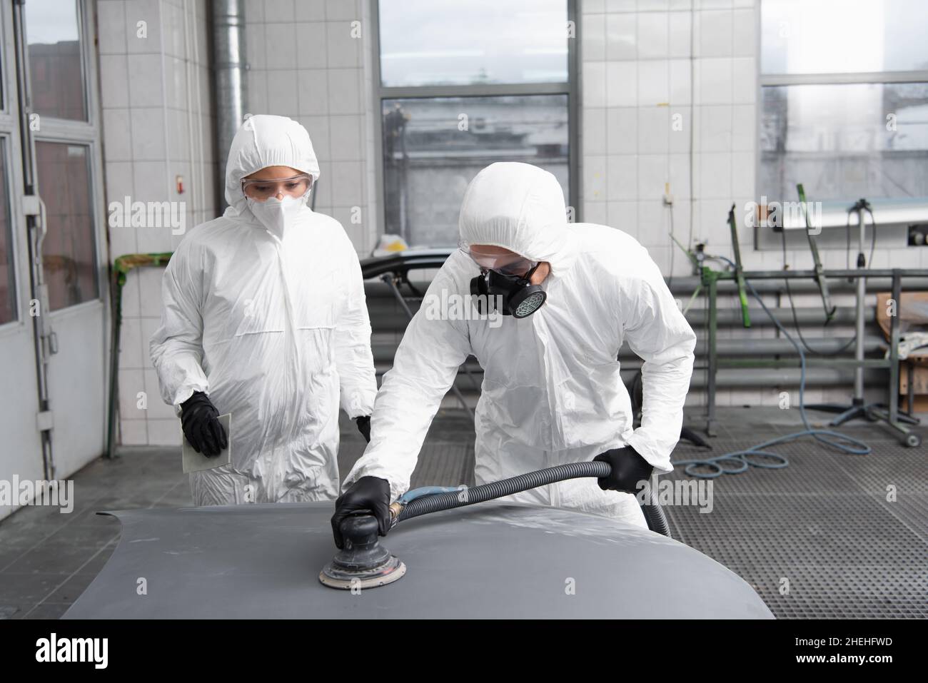 Workman in hazmat suit and respirator polishing auto near colleague in garage Stock Photo