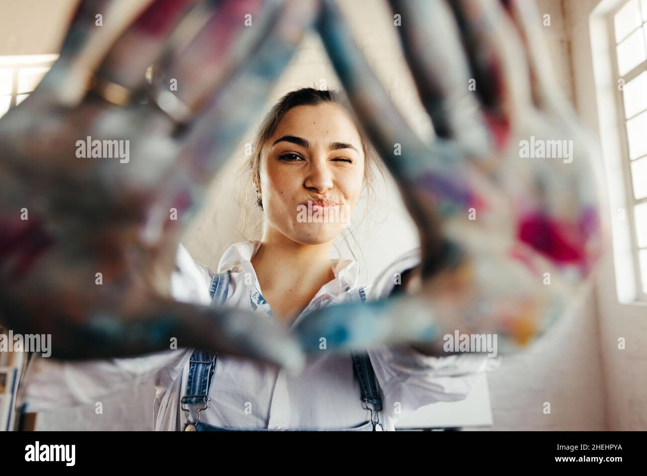 Carefree young painter framing her face with her hands. Happy female artist winking at the camera while standing in her art studio. Creative young wom Stock Photo
