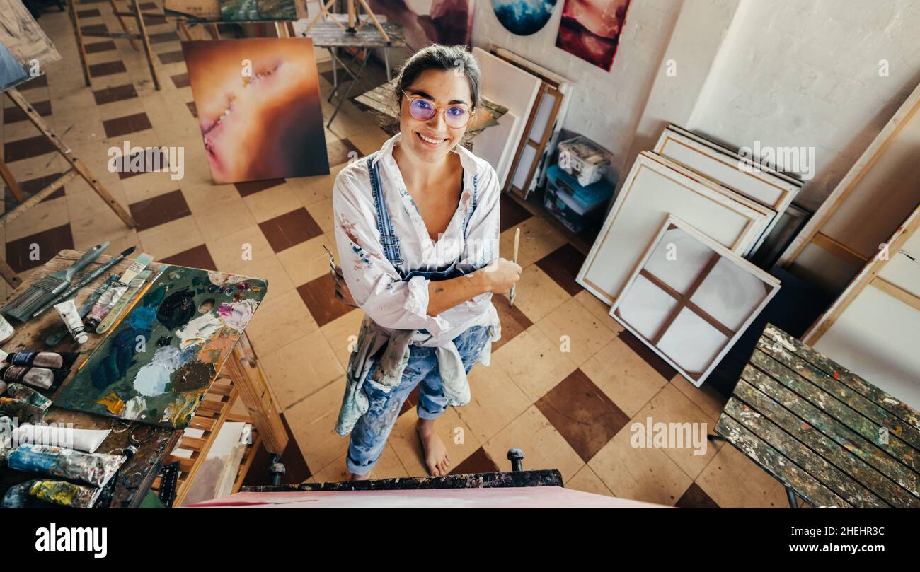 Happy female artist smiling in her art studio. Imaginative young painter looking at the camera while standing with her arms crossed. Creative young wo Stock Photo