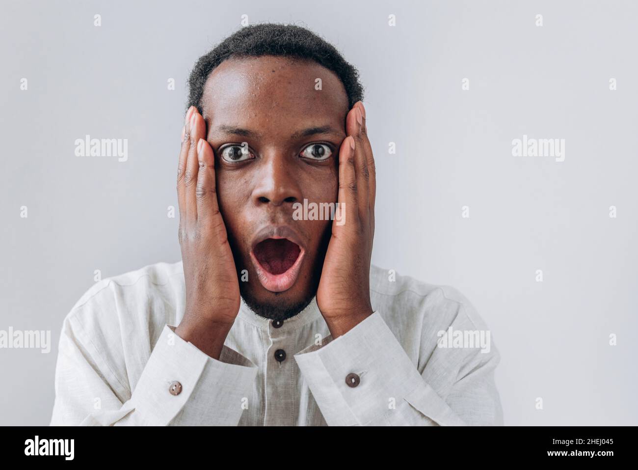 Portrait of funny shocked African-American man in white shirt holding cheeks standing on light grey background close view Stock Photo