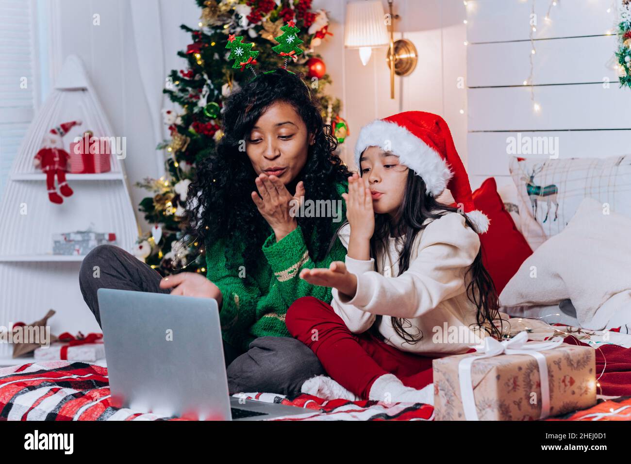 African American woman and brunette daughter in Santa hat sit among Christmas decorations and blow kisses chatting on video call via laptop Stock Photo