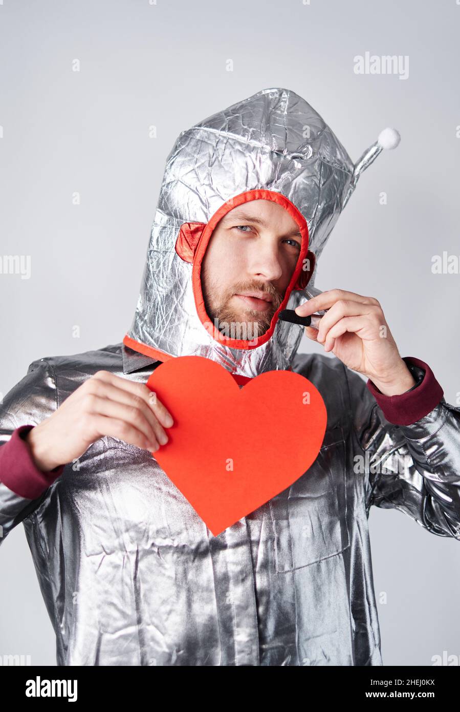 Male in spacesuit with red paper heart talking to somebody greeting with St. Valentine's Day. Romance, relationships concept. Young bearded astronaut man standing by the wall. High quality image Stock Photo