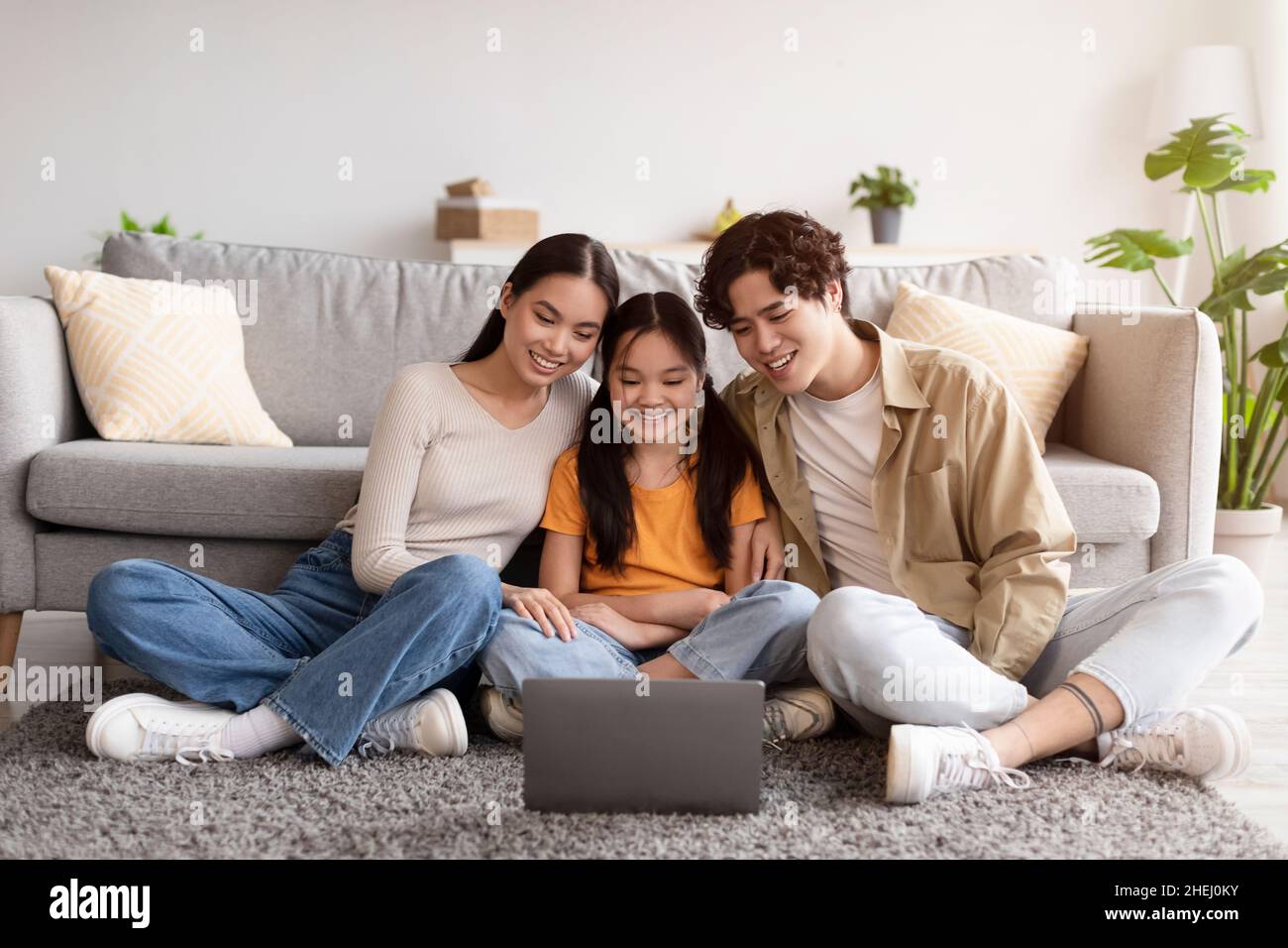 Satisfied millennial japanese mom, dad and teen daughter sitting on floor and watching video on laptop Stock Photo