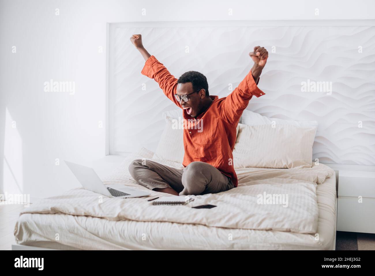 Joyful African-American guy in casual clothes and glasses puts hands up sitting near open laptop on comfortable bed at home Stock Photo