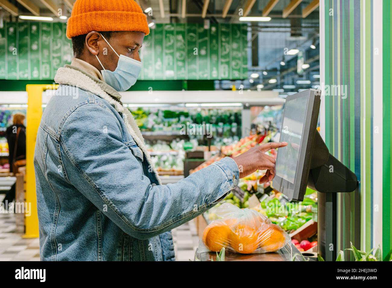 Stylish African-American guy in warm jacket with disposable mask weighs fresh oranges with digital scale in light supermarket Stock Photo
