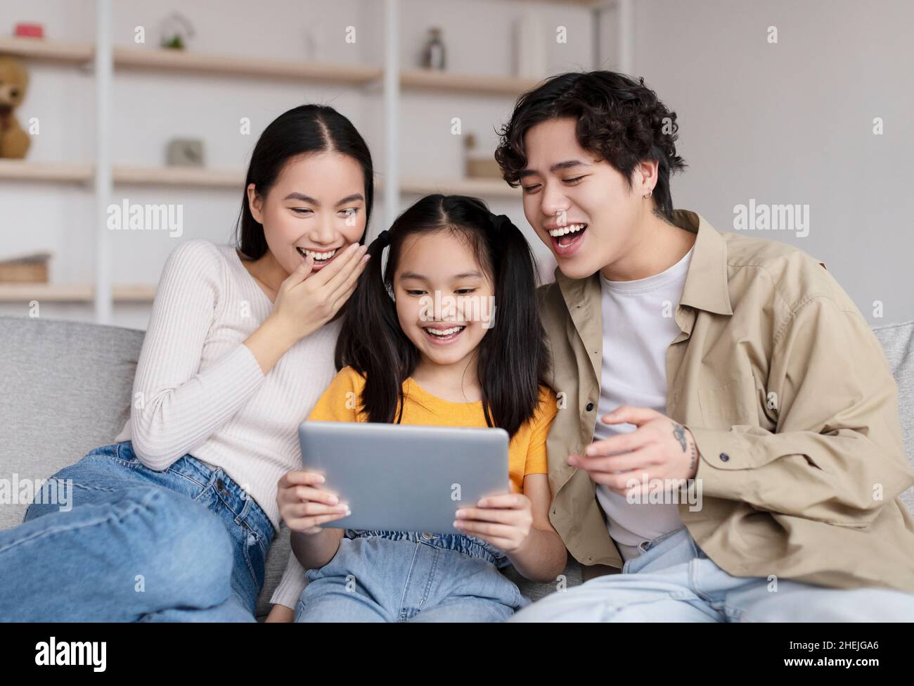 Laughing young asian husband, wife and teen daughter watching funny video on tablet, surfing in internet Stock Photo