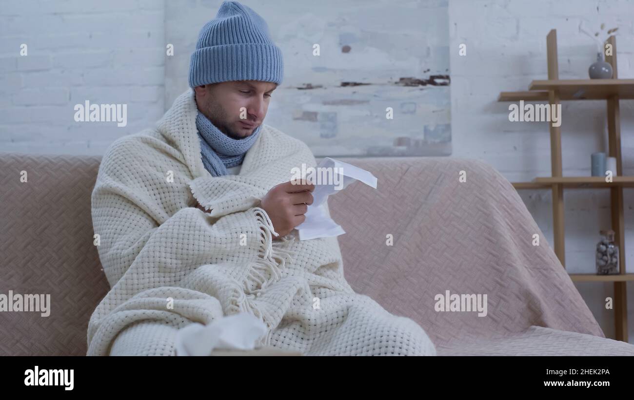 ill man in warm beanie and blanket reading medication instruction on couch at home Stock Photo