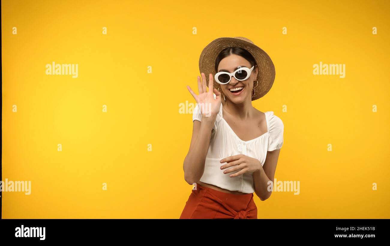 Smiling model in sunglasses and straw hat waving hand isolated on yellow Stock Photo