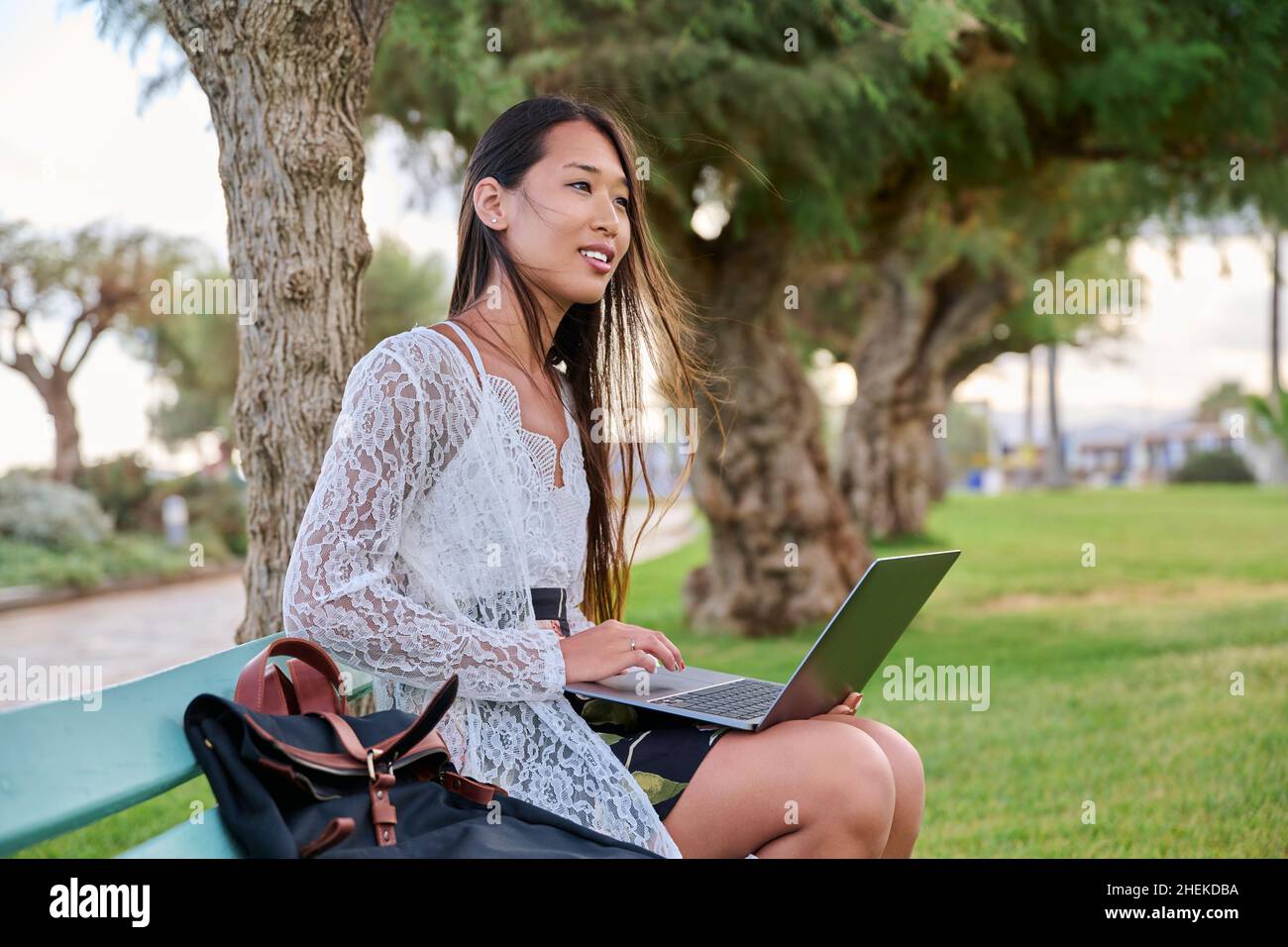 Beautiful young Asian woman using laptop sitting on an outdoor bench ...