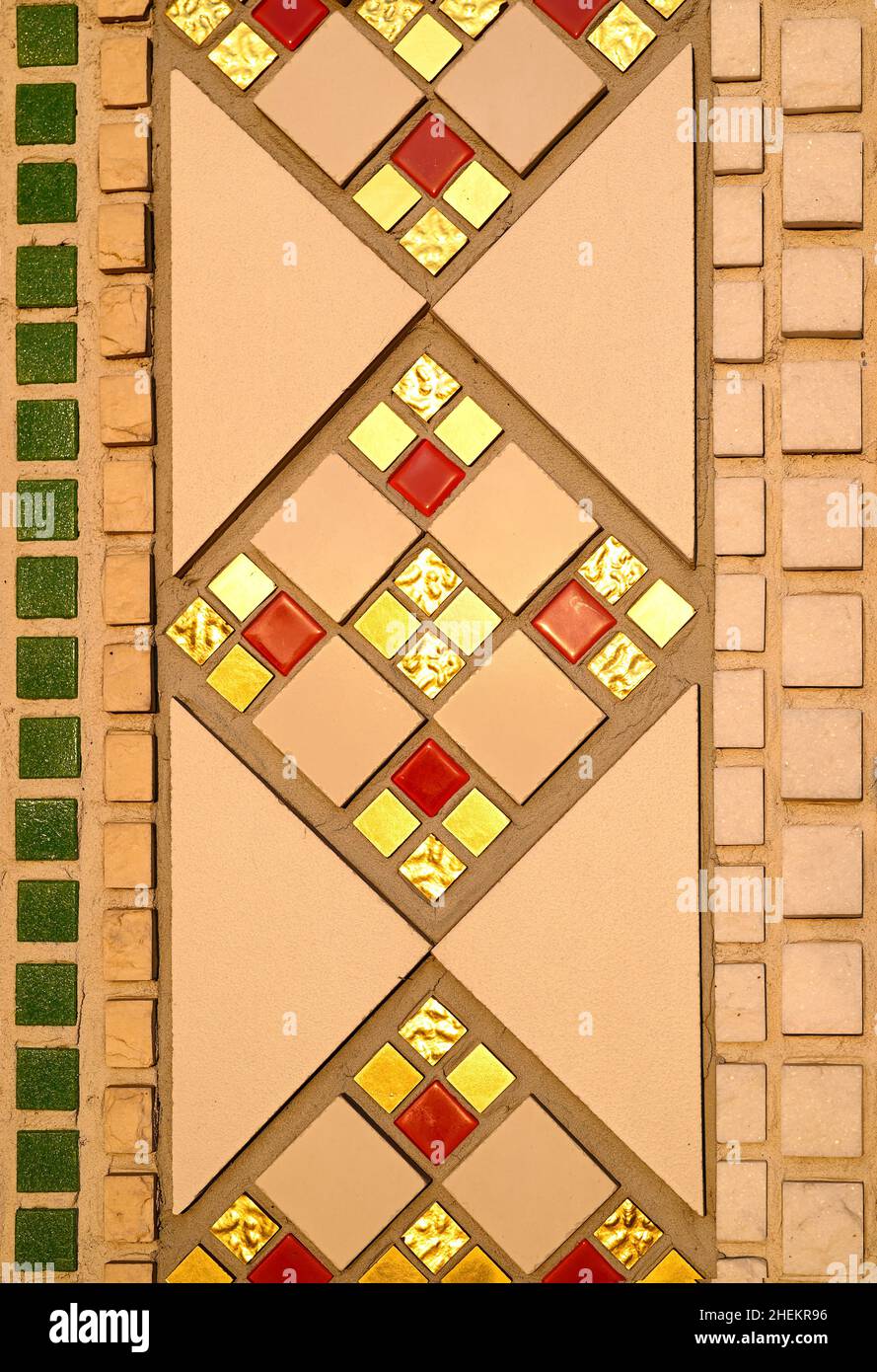 A fragment of a geometric repeating pattern from a ceramic mosaic on the wall of a Catholic church. Gold, red, beige and green mosaic tile. Stock Photo