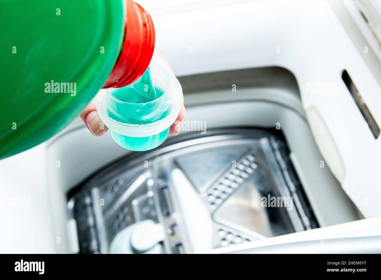 Liquid detergent from a green bottle is poured into the washing machine. Preparation for washing, measuring cup for washing powder. Stock Photo