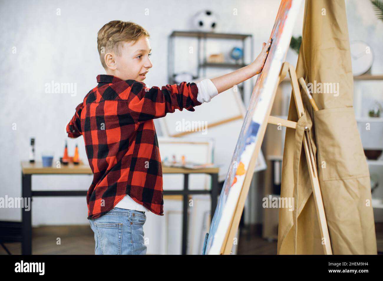 Inspired caucasian boy creating his masterpiece on easel using paints and hands. Little student enjoying art lesson at modern school. Stock Photo