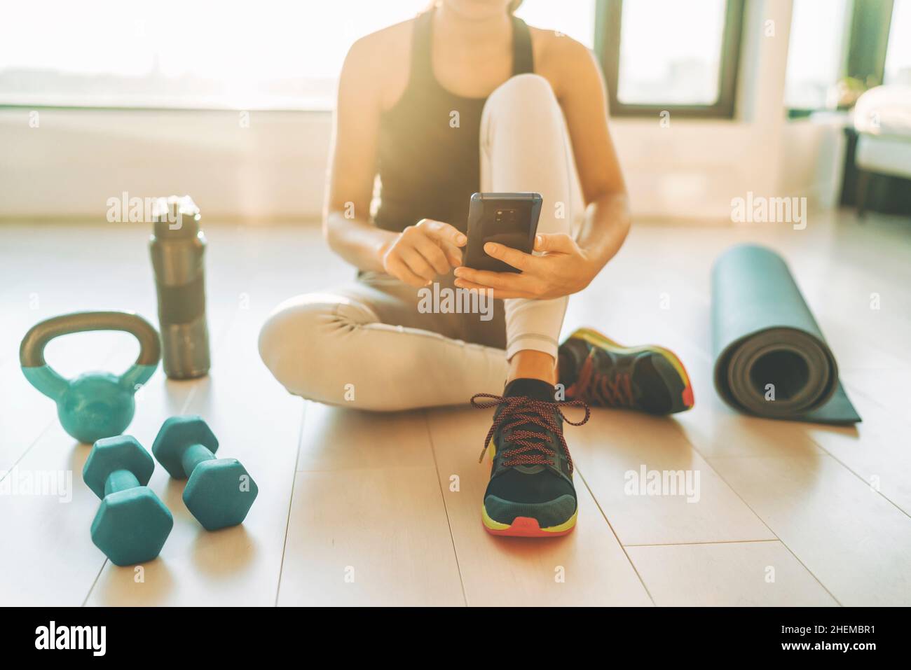 Fitness app on mobile phone for exercise at home. Woman using tech device during strength training workout with dumbbells and kettlebell Stock Photo