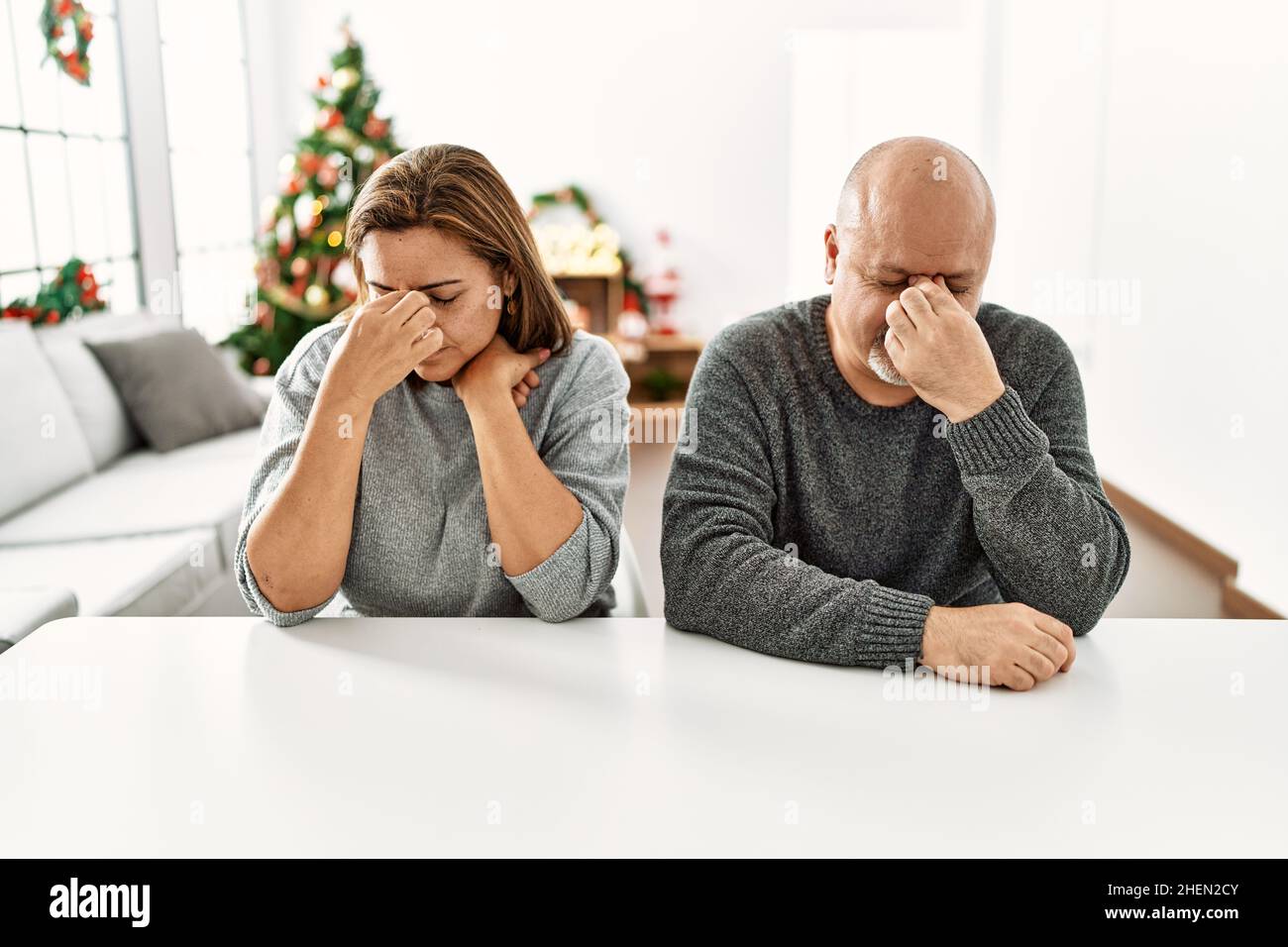 Middle age hispanic couple sitting on the table by christmas tree tired