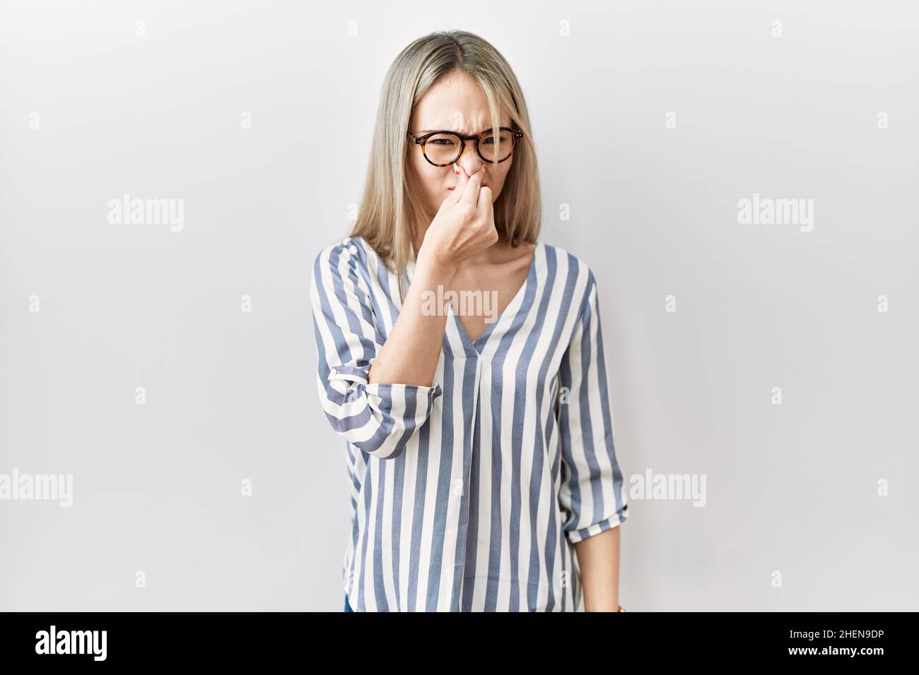 Asian young woman wearing casual clothes and glasses smelling something stinky and disgusting, intolerable smell, holding breath with fingers on nose. Stock Photo