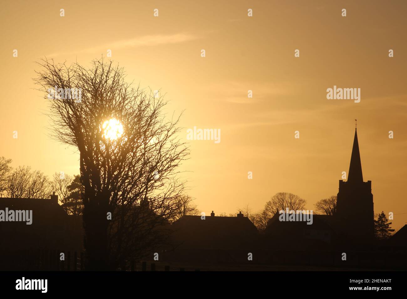 Silhouettes of houses and St John the Baptist C. of E. church in Pilling, Preston, Lancashire, England at sunset with the sun behind bare tree. Stock Photo