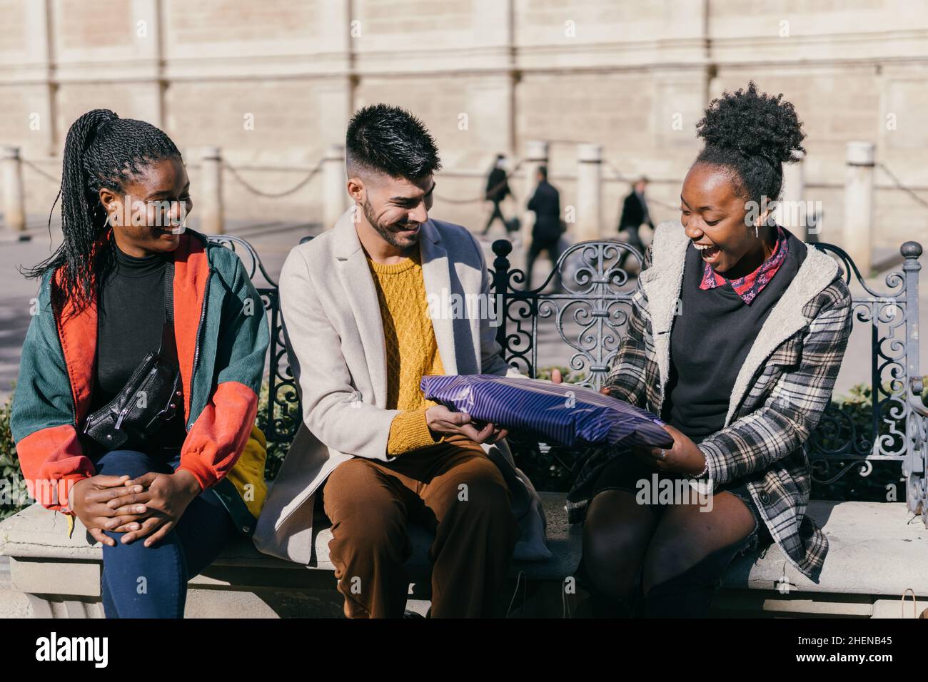 Three friends exchanging presents while sitting on a bench outdoors Stock Photo