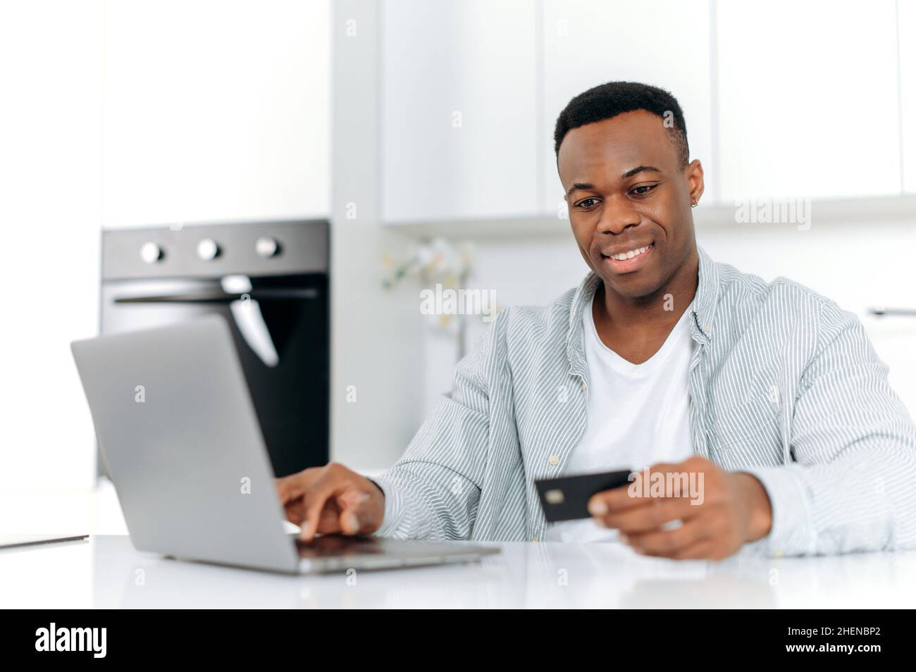 Joyful black man sits on kitchen at home, dressed in casual wear, using laptop and credit card for online shopping, is entering data for payment, online transaction, order home delivery, smiles Stock Photo