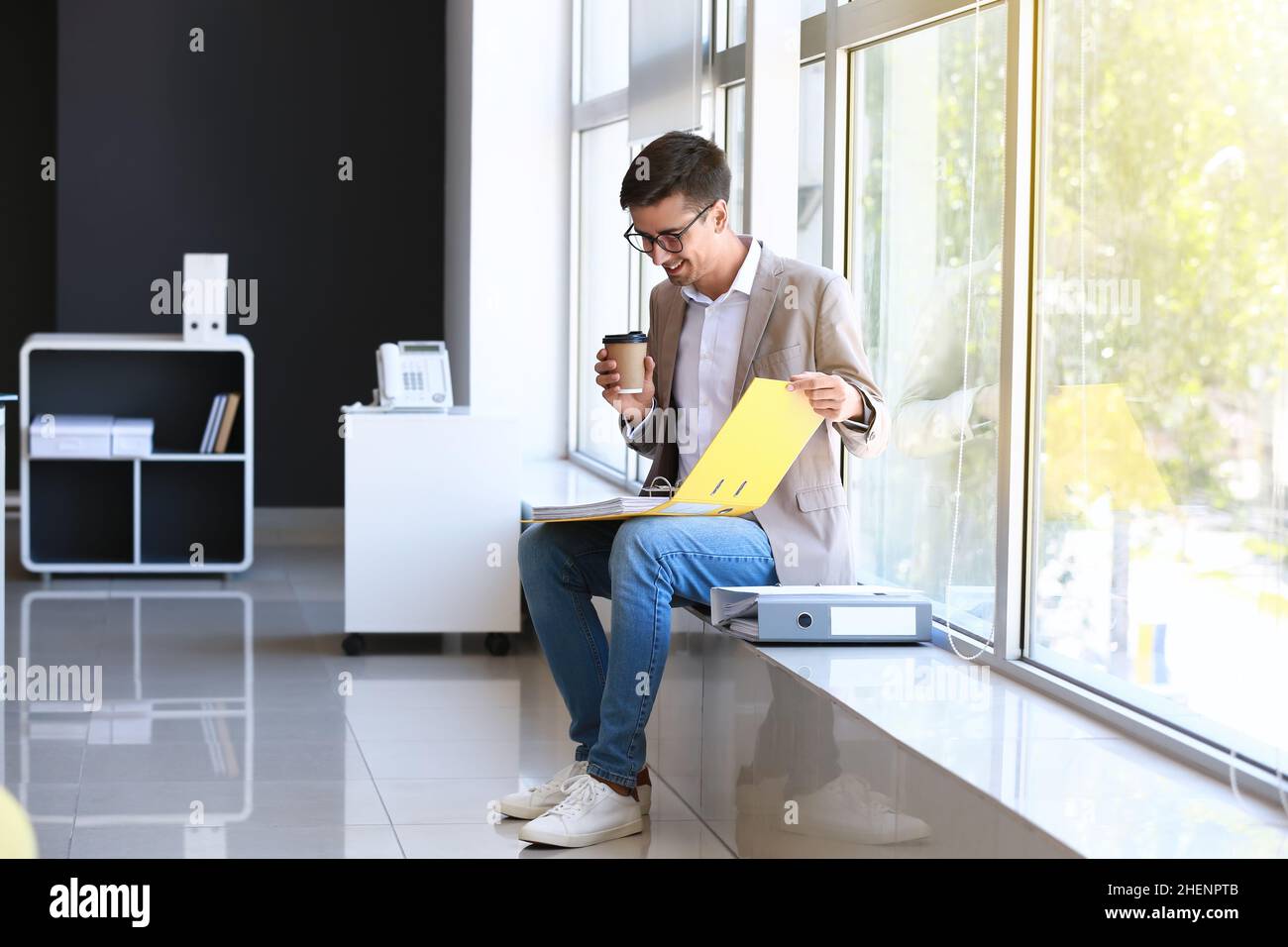 Young man with open folder and cup of coffee sitting on windowsill in ...
