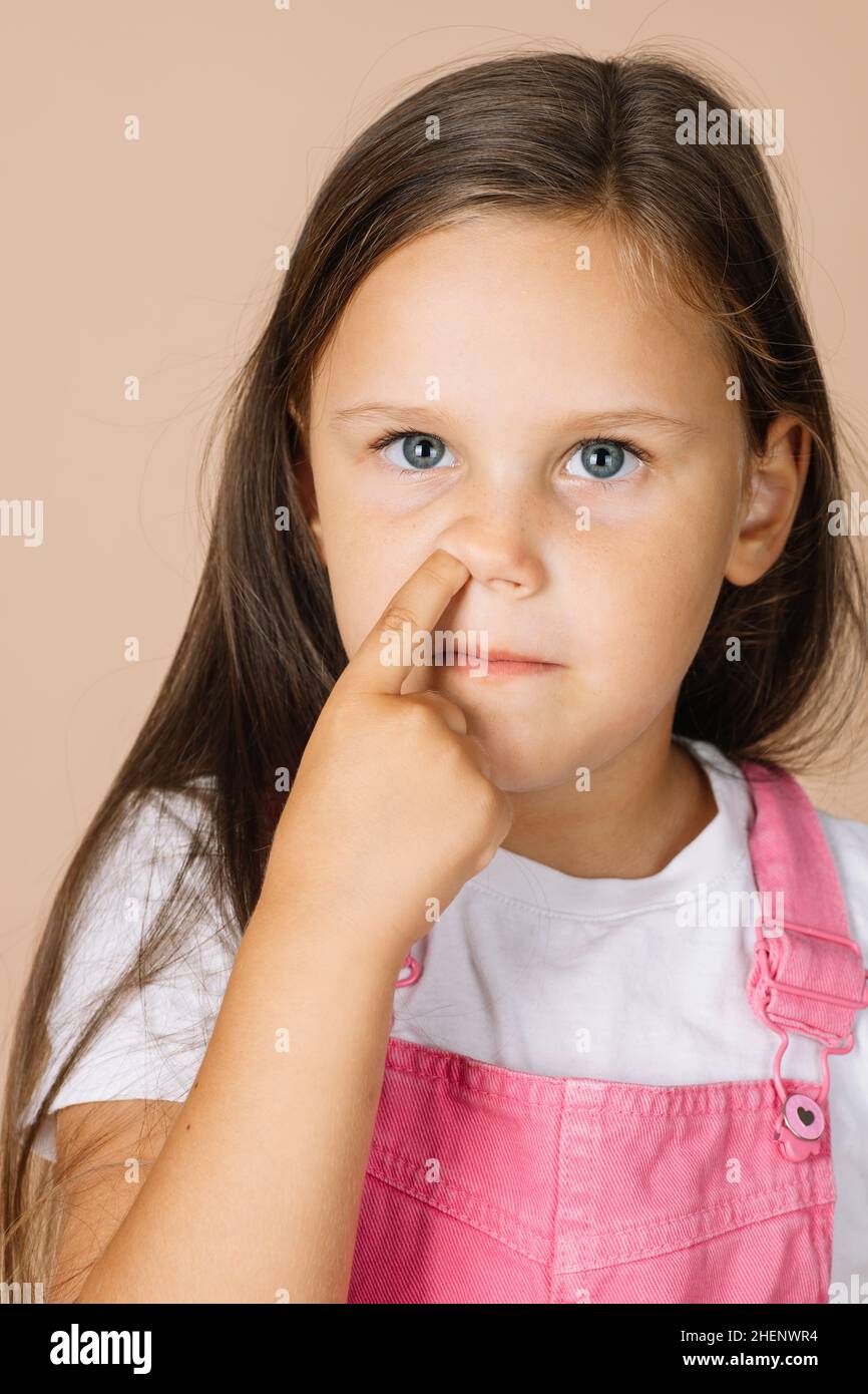Portrait photo of female kid picking nose with point finger with shining eyes looking upward wearing bright pink jumpsuit and white t-shirt on beige Stock Photo