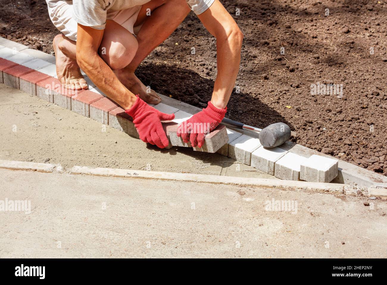A young worker lays paving slabs on a bright sunny summer day on the prepared sandy base of a park path under construction. Copy space. Stock Photo