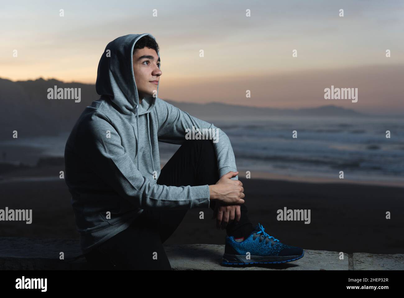 Portrait of handsome hispanic teenager resting and sitting at sunset, in front of the beach Stock Photo