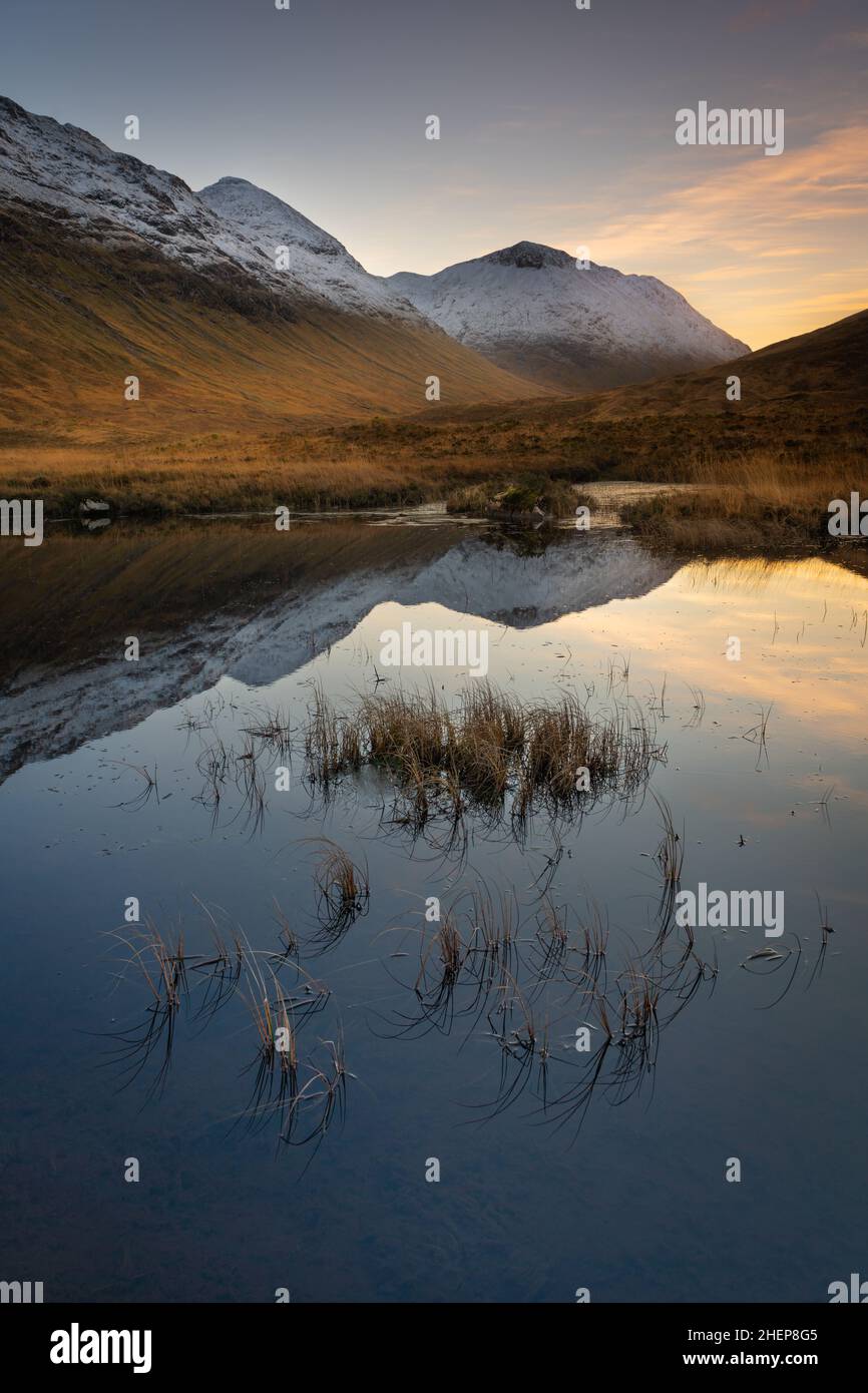 Glencoe Scotland, Lochan na Fola, standing in highland marshes & deep grass, snow capped mountains left & right & a mirror reflection on the water. Stock Photo