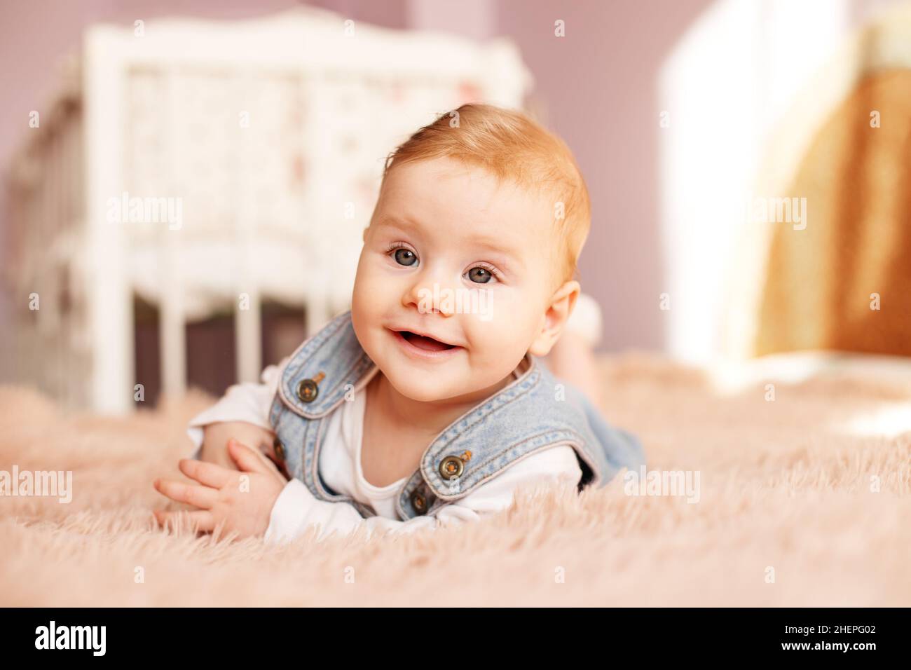 Newborn 5 months old lies on bed on stomach. Baby girl smiles and plays in room. Denim sundress. Soft bedspread. Stock Photo