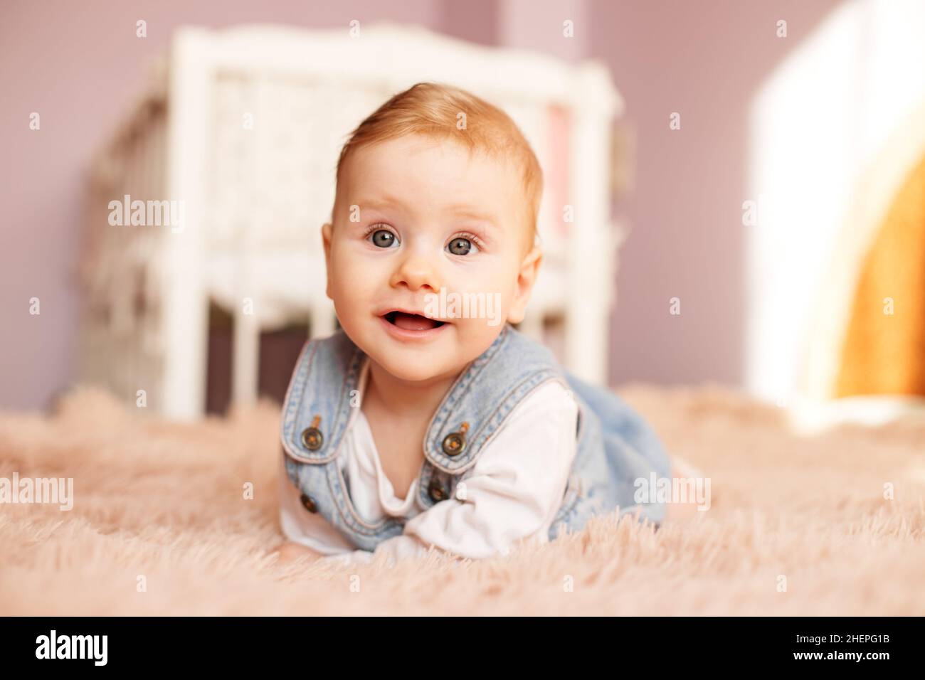 Newborn 5 months old lies on bed on stomach. Baby girl smiles and plays in room. Denim sundress. Soft bedspread. Stock Photo