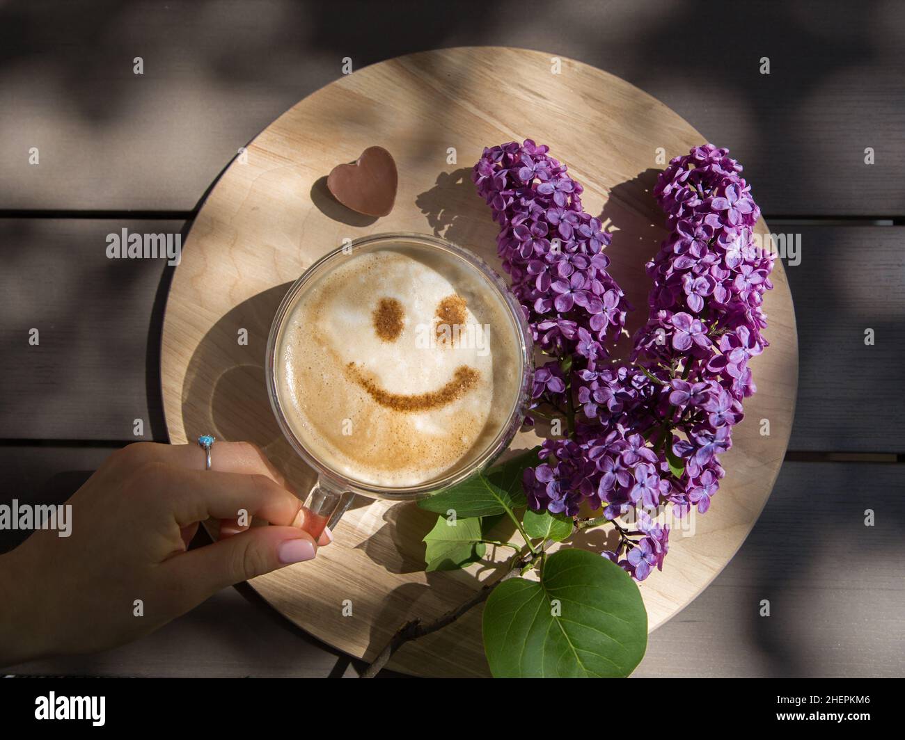 Lilac, a female hand near a cup of coffee with a smile from cinnamon on milk foam on a round wooden stand. View from above. Positive thinking, good mo Stock Photo