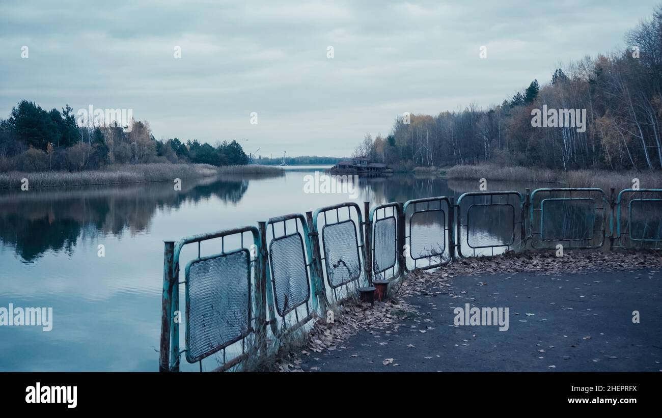 pripyat river and embankment with rusty fence under grey cloudy sky in chernobyl exclusion zone Stock Photo