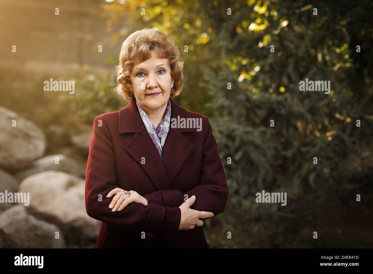 Beautiful adult caucasian woman with short blond hair stands with crossed arms in the park in a burgundy coat and a scarf. High quality photo Stock Photo