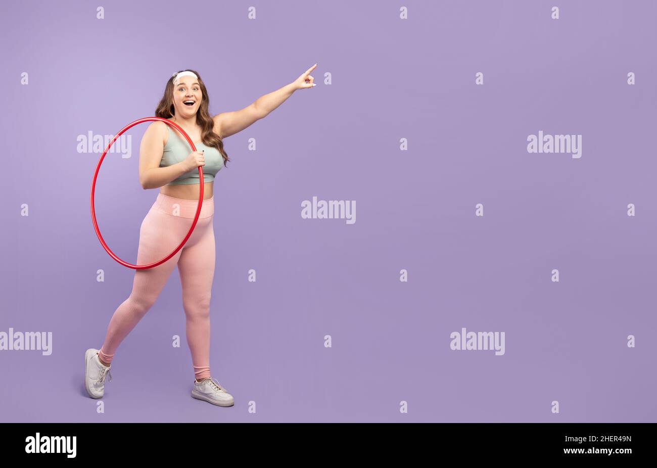 Happy millennial european plus size woman in sports uniform with hula hoop pointing to blank space Stock Photo