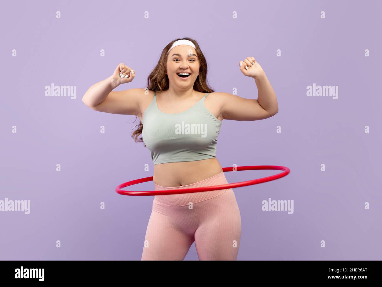 Cheerful millennial european plus size fit lady in sports uniform twirls hula hoop isolated on purple background Stock Photo