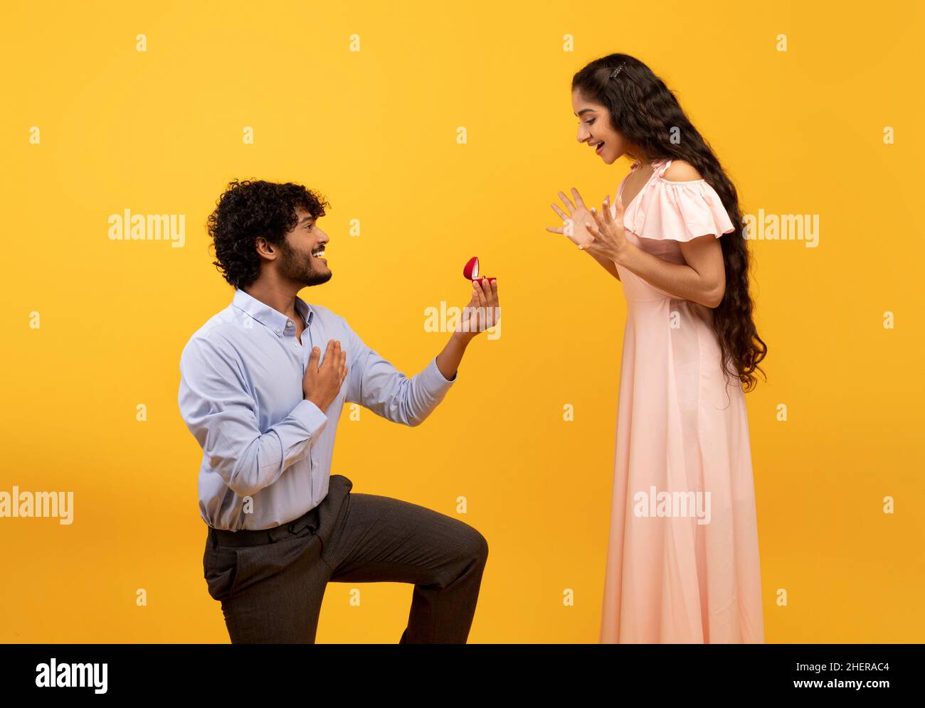 Loving indian man standing on one knee and offering engagement ring to his beloved woman on yellow background Stock Photo