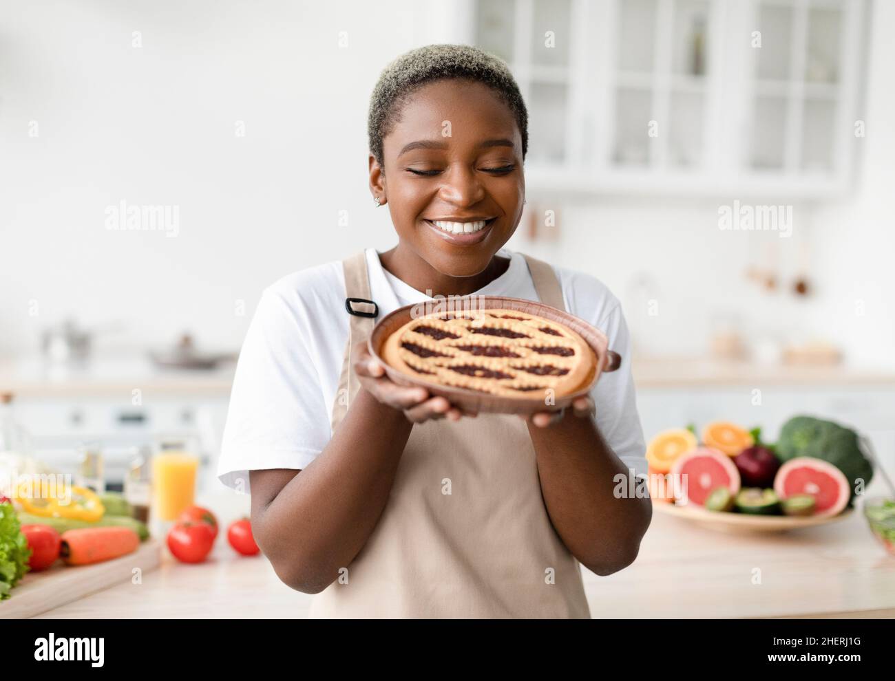 Smiling pretty young african american woman in apron sniffs smell of fresh pie in scandinavian kitchen interior Stock Photo