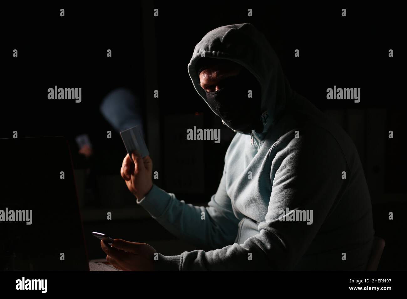 Hacker in dark jacket looking at laptop screen in dark room is holding bank card Stock Photo