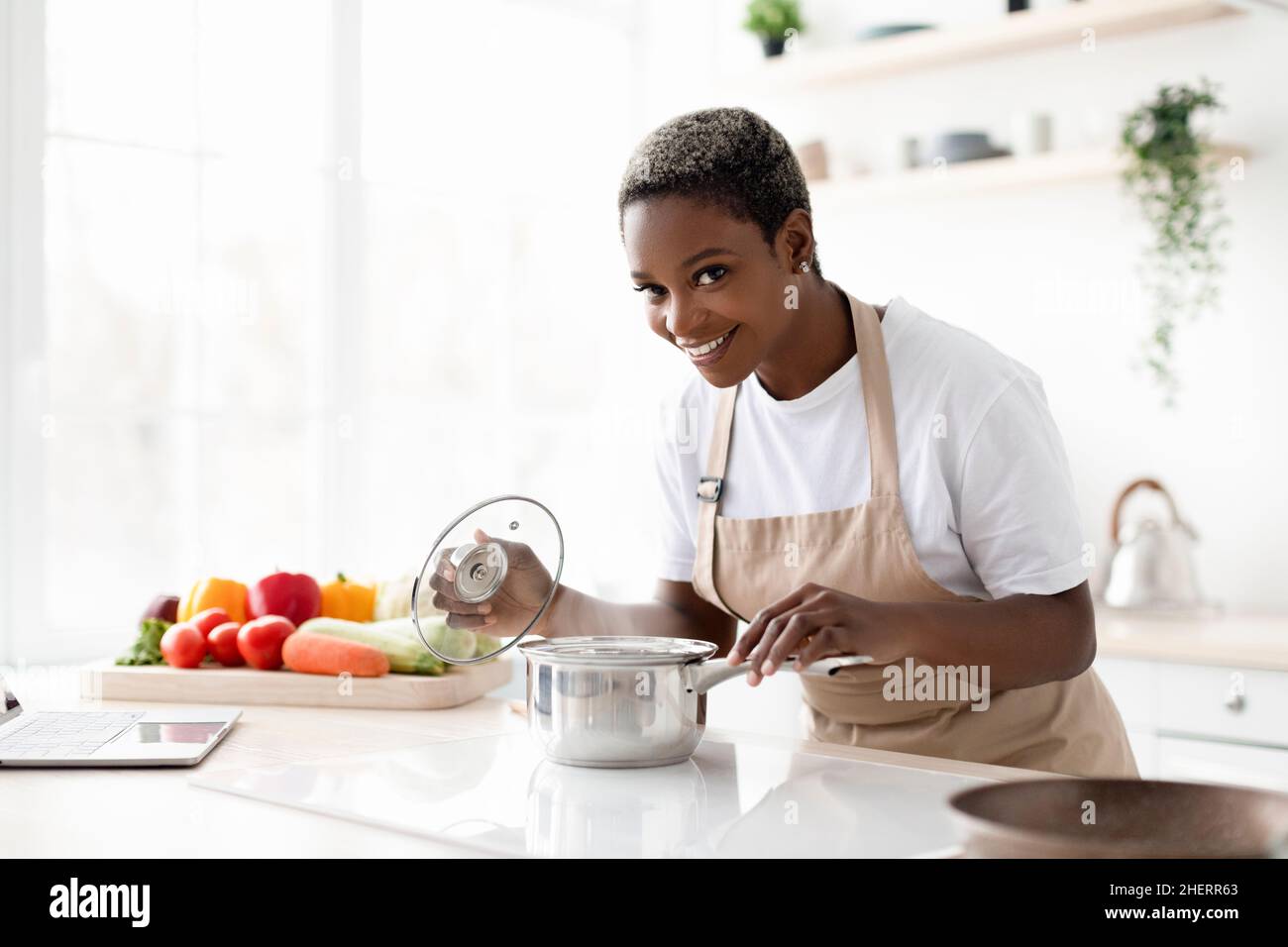 Smiling young african american woman in apron prepare eat and smell dish in minimalist kitchen interior Stock Photo