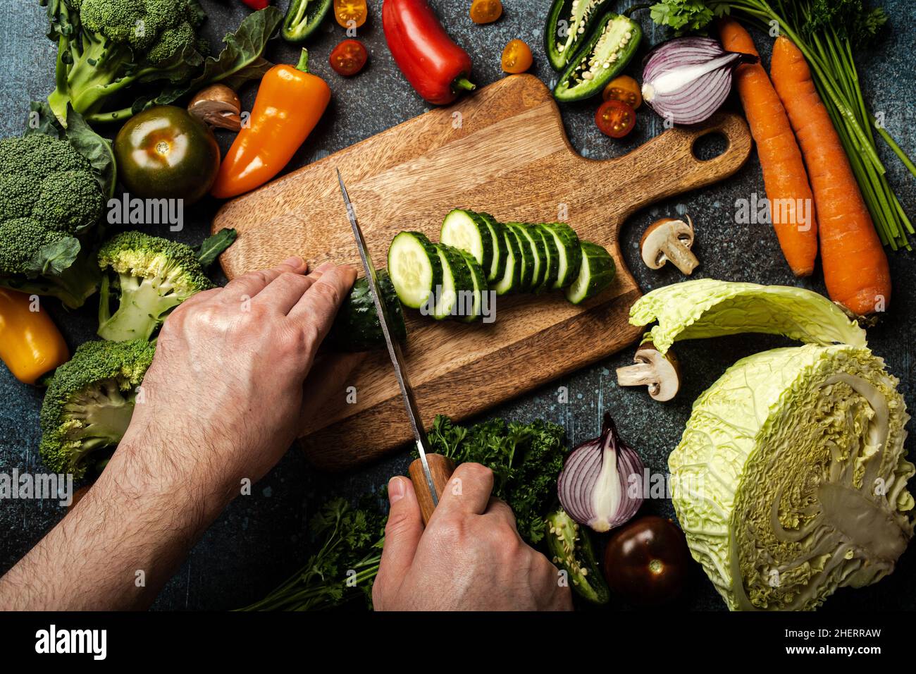 Man hands chopping cucumber on wooden cutting board with knife and assorted fresh vegetables Stock Photo
