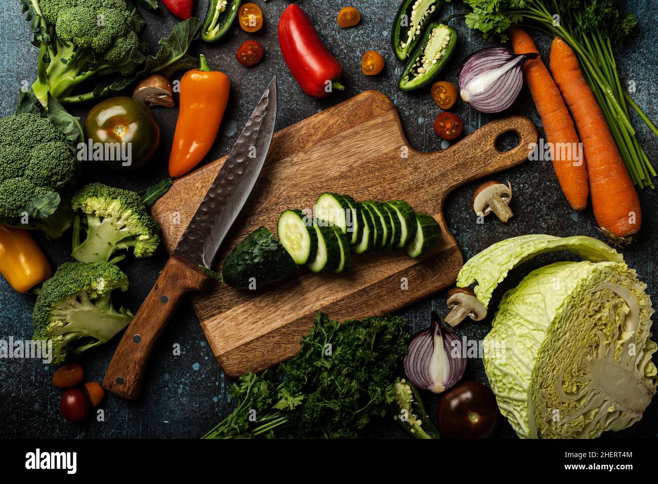 Chopped cucumber on wooden cutting board with knife and assorted fresh vegetables Stock Photo