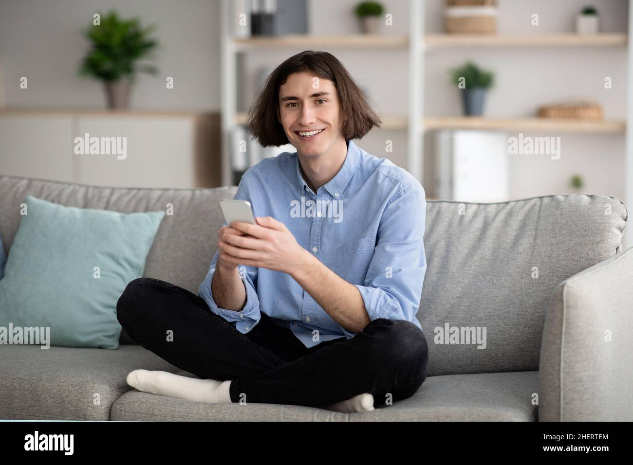 Enjoying weekend. Happy young guy using smartphone while sitting cross legged on couch at home, copy space Stock Photo