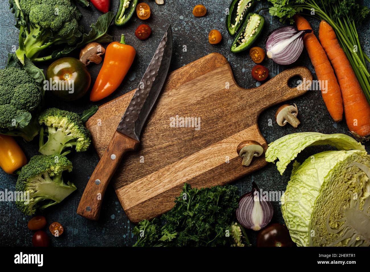 Assorted fresh vegetables on rustic concrete table with wooden cutting board in center and kitchen knife Stock Photo