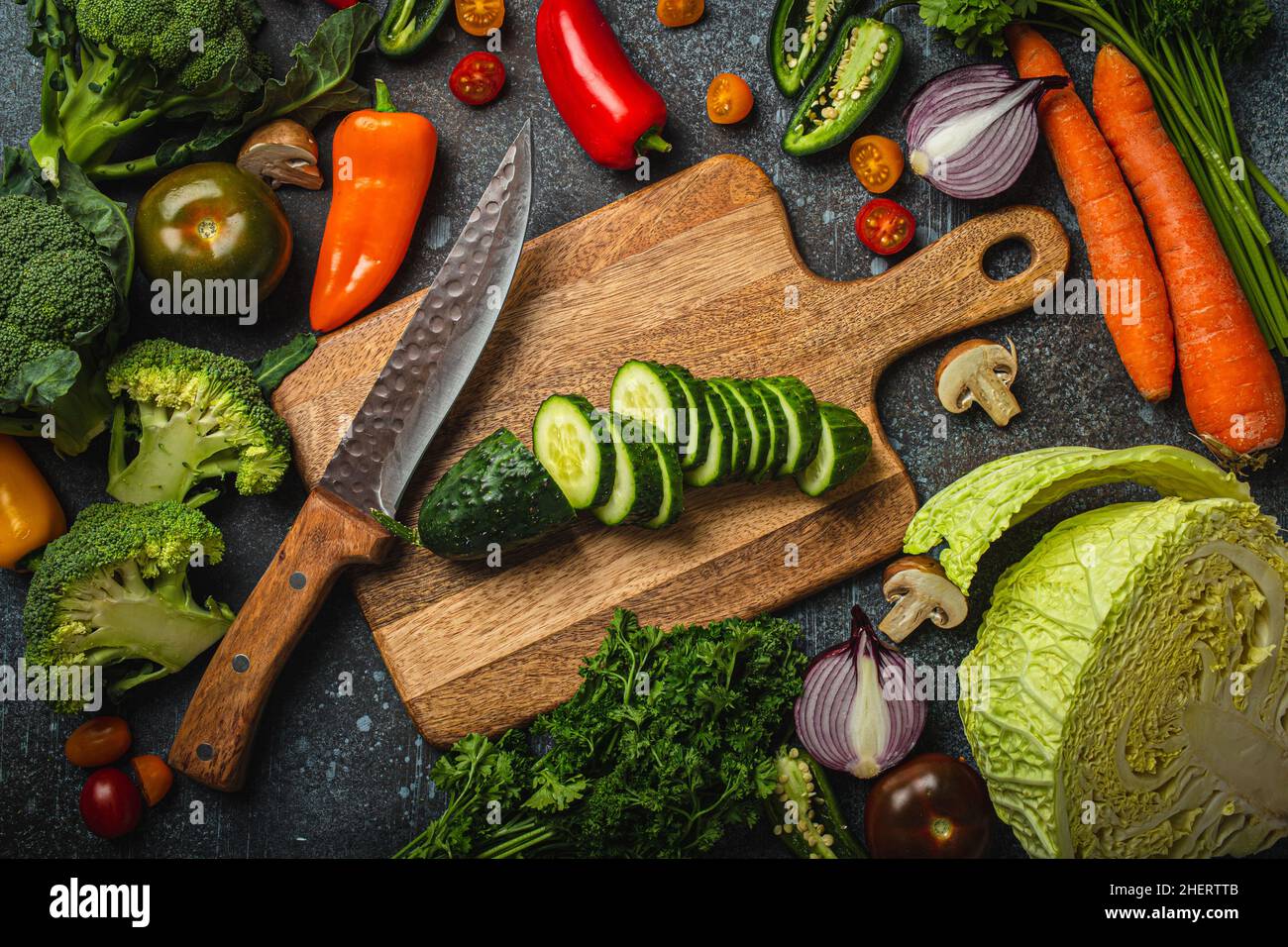 Chopped cucumber on wooden cutting board with knife and assorted fresh vegetables Stock Photo