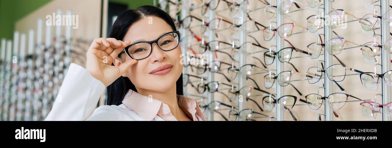 happy asian oculist looking at camera and touching eyeglasses in optics ...